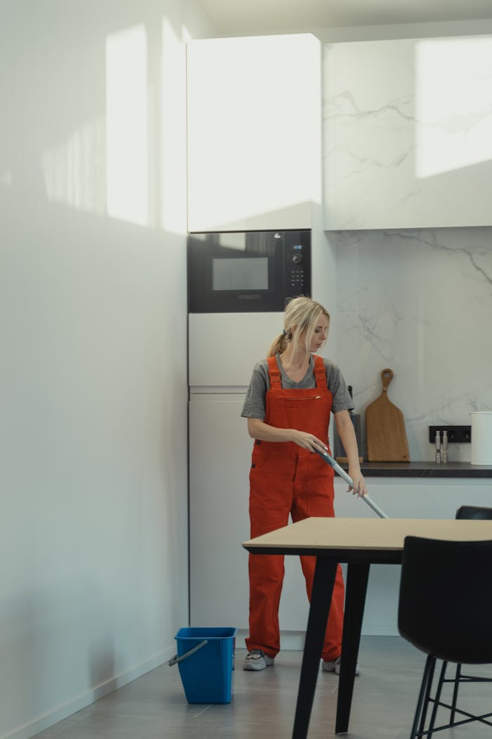 A woman in red overalls mopping a kitchen floor under bright daylight, depicting housekeeping chores.